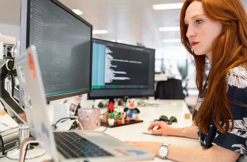 A focused view of a woman working on a computer, screen displayed on dual monitors in an office environment.