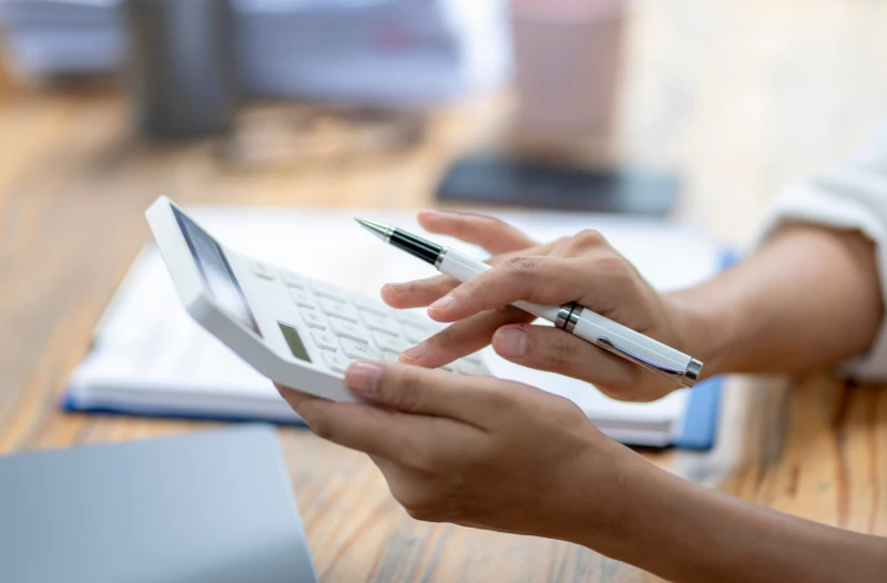 A close-up of hands holding a pen and calculator, with financial documents in the background.